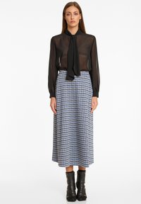 Schwarze durchsichtige Bluse mit Bindeband am Hals, kombiniert mit einem blau-weißen karierten Midi-Rock. Das Model trägt schwarze Stiefeletten.