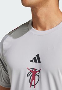 Gray athletic shirt with a round neckline. Features black Adidas logo and red graphic of a figure with outstretched arms. Smooth texture.