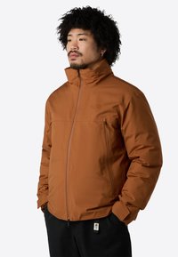 Braune isolierte Jacke mit hohem Kragen, Reißverschluss und Seitentaschen. Glatte Textur, minimale Markenkennzeichnung auf der Brust.
