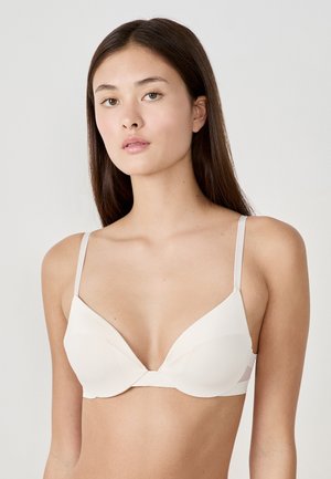 Soutien-gorge nude clair avec un décolleté plongeant, en tissu lisse, avec des bretelles fines et ajustables, et un détail de couture subtil au centre.