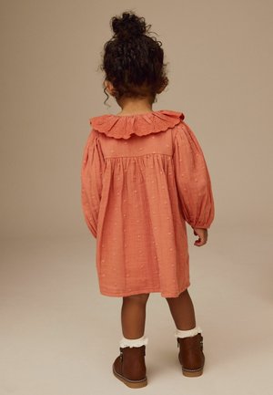 Jeune enfant aux cheveux bouclés coiffés en chignon haut, portant une robe corail texturée avec un col à volants et des bottes marron avec des chaussettes blanches en dentelle, debout.