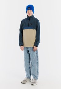 Fleecejacke in Marineblau und Beige mit Reißverschlusskragen, versehen mit blauem Logo. Blauer Strickmütze. Hellblaue Jeans und graue Sneaker runden den Look ab.