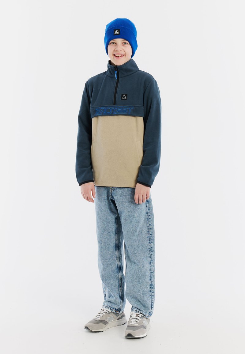 Fleecejacke in Marineblau und Beige mit Reißverschlusskragen, versehen mit blauem Logo. Blauer Strickmütze. Hellblaue Jeans und graue Sneaker runden den Look ab.