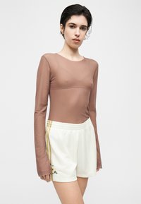 Νεαρό άτομο με κοντά σκούρα μαλλιά φορά διάφανο καφέ μακρυμάνικο τοπ και λευκά αθλητικά σορτς με πλευρικές ρίγες και μικρή κεντημένη λουλουδένια λεπτομέρεια.