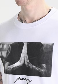 White cotton t-shirt featuring a black and white photographic print of hands in a prayer position with tattoos, and the word “pray” in black below.