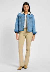 Chaqueta de denim con cuello de sherpa, color azul claro, dos bolsillos en el pecho, cierre frontal de botones, combinada con pantalones acampanados beige y botas de tobillo beige.