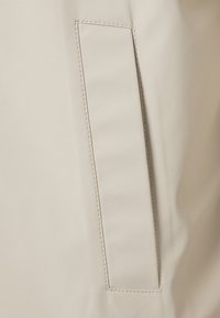 Tissu de veste beige avec une texture lisse, comprenant une poche plaquée rectangulaire et des coutures contrastantes le long des bords.