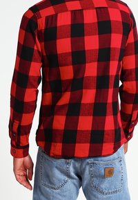 Camisa de franela a cuadros rojo y negro, con mangas largas, puños abotonados y un detalle de yugo en la parte de atrás, combinada con jeans de color azul claro.