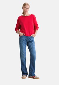 Roter Pullover mit langen Ärmeln, kombiniert mit blauen geraden Jeans. Das Modell trägt lässige Schuhe mit einer strukturierten Oberfläche und Gummisohlen.