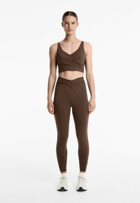 Brun kort sportbehå med tvinnad framsida, tillsammans med högmidjade leggings i matchande nyans. Skor: vita sportskor.