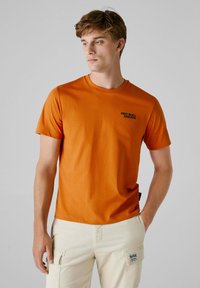 Kortærmet orange t-shirt, lavet af bomuldsmateriale, rund halsudskæring, med teksten "RED BULL RACING" i sort på venstre brystområde.