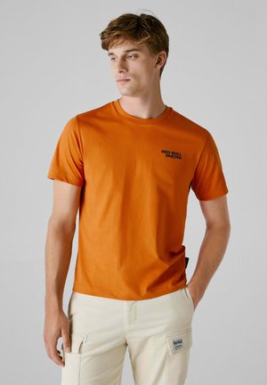 Camiseta de manga corta color naranja, de material de algodón, con cuello redondo, que presenta el texto "RED BULL RACING" en negro en la zona del pecho izquierdo.