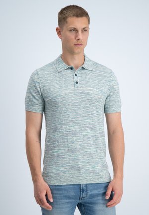 Uomo con polo a maniche corte in maglia mélange azzurro chiaro e jeans blu, in piedi davanti a uno sfondo chiaro e uniforme.