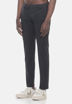 Uomo con pantaloni slim neri e sneakers bianchi, in piedi davanti a uno sfondo chiaro e semplice.