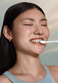 Brosse à dents électrique avec un manche blanc et des poils bleus, tenue près de la bouche d'une femme. Elle a de longs cheveux bruns et porte un haut bleu clair.