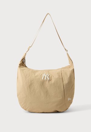 Beżowa torba na ramię z regulowanym paskiem, z białym haftowanym logo New York Yankees z przodu i małym logo marki w pobliżu dolnej strony.