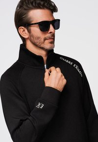Sudadera negra con cremallera y cuello alto, con letras blancas en el pecho y la manga. El modelo lleva gafas de sol oscuras.