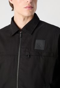 Jachetă neagră cu fermoar, fabricată dintr-un material neted, având un guler în vârf, buzunar frontal și o etichetă cu logo. Design minimalist cu fermoar rezistent.