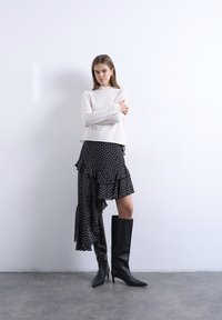 Γυναίκα που στέκεται με σταυρωμένα χέρια, φορώντας κρέμ πουλόβερ, ασύμμετρη μαύρη φούστα με πουά και μαύρες ψηλοτάκουνες μπότες μέχρι τα γόνατα, μπροστά σε λευκό τοίχο.