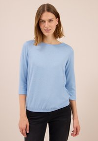 Frau mit schulterlangem Haar, die ein hellblaues 3/4-Ärmel-Oberteil und schwarze Hose trägt, steht vor einem beigen Hintergrund.
