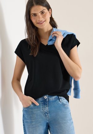 Ung kvinde med langt brunt hår iført en sort V-hals T-shirt og blå jeans, der holder en lyseblå trøje over skulderen.