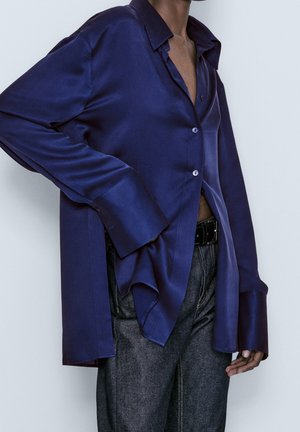 Satin, dybblå skjorte med knapper, løs pasform, overdimensionerede manchetter og delt kant, båret over mørke denimbukser.