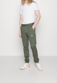 Kapsáčové kalhoty z bavlny olivově zelené barvy, s boční kapsou a klasickým střihem. Kombinované s bílým tričkem a bílými teniskami s oranžovými detaily.