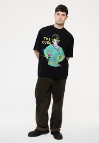 Black oversized t-shirt featuring colorful graphic print of a person with the text "THE CURE." Paired with brown corduroy pants and black shoes.