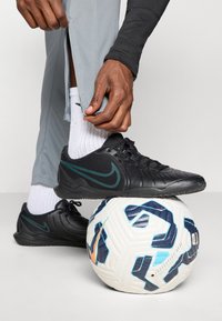 Black athletic shoes with textured surface and green accents, positioned on a white soccer ball with blue and orange designs. Gray athletic pants.