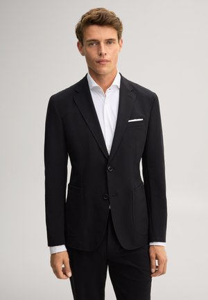Giovane uomo con abito nero, camicia bianca e fazzoletto da taschino, in piedi davanti a uno sfondo grigio chiaro uniforme.