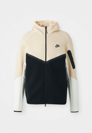 Sudadera con cremallera de Nike que presenta un diseño bicolor en beige, negro y blanco. El material es suave, cuenta con capucha y un bolsillo con cremallera en la manga.