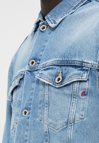 Φωτεινό μπλε τζιν μπουφάν με κουμπιά μπροστά, δύο τσέπες στο στήθος, ραφές σε αντίθεση και μικρό κόκκινο λογότυπο "R" στην δεξιά τσέπη.