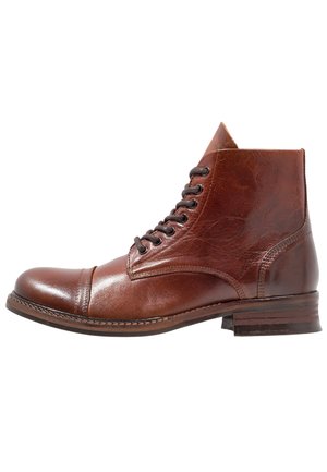 Ruskea nahkainen nauhollinen nilkkuri kärkipäällisellä ja pinosalpa-korolla, esitetty ulkokuorelta valkoista taustaa vasten.