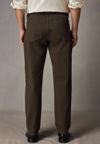 Bruine, rechte broek met een gladde textuur, vijf zakken en zichtbare stiksels, gedragen met zwarte schoenen.