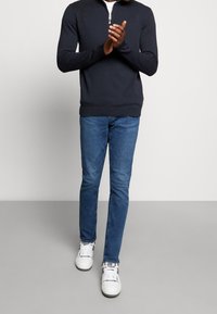 Maglione navy a collo zip abbinato a jeans blu e sneakers bianche. Il maglione presenta polsini e orlo a coste, e una texture liscia.
