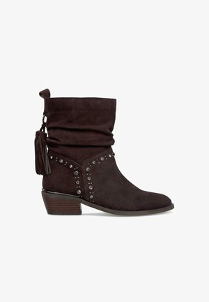 Stivale alla caviglia in suede marrone con design scivolato, accenti di borchie e dettaglio con nappina; presenta un tacco basso impilato e una suola liscia.