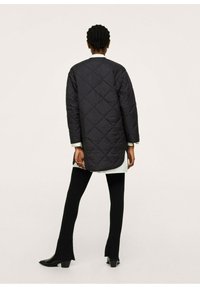 Cappotto lungo ondulato nero, visto da dietro. Presenta polsini a coste ed è abbinato a pantaloni neri attillati e stivali con tacco.
