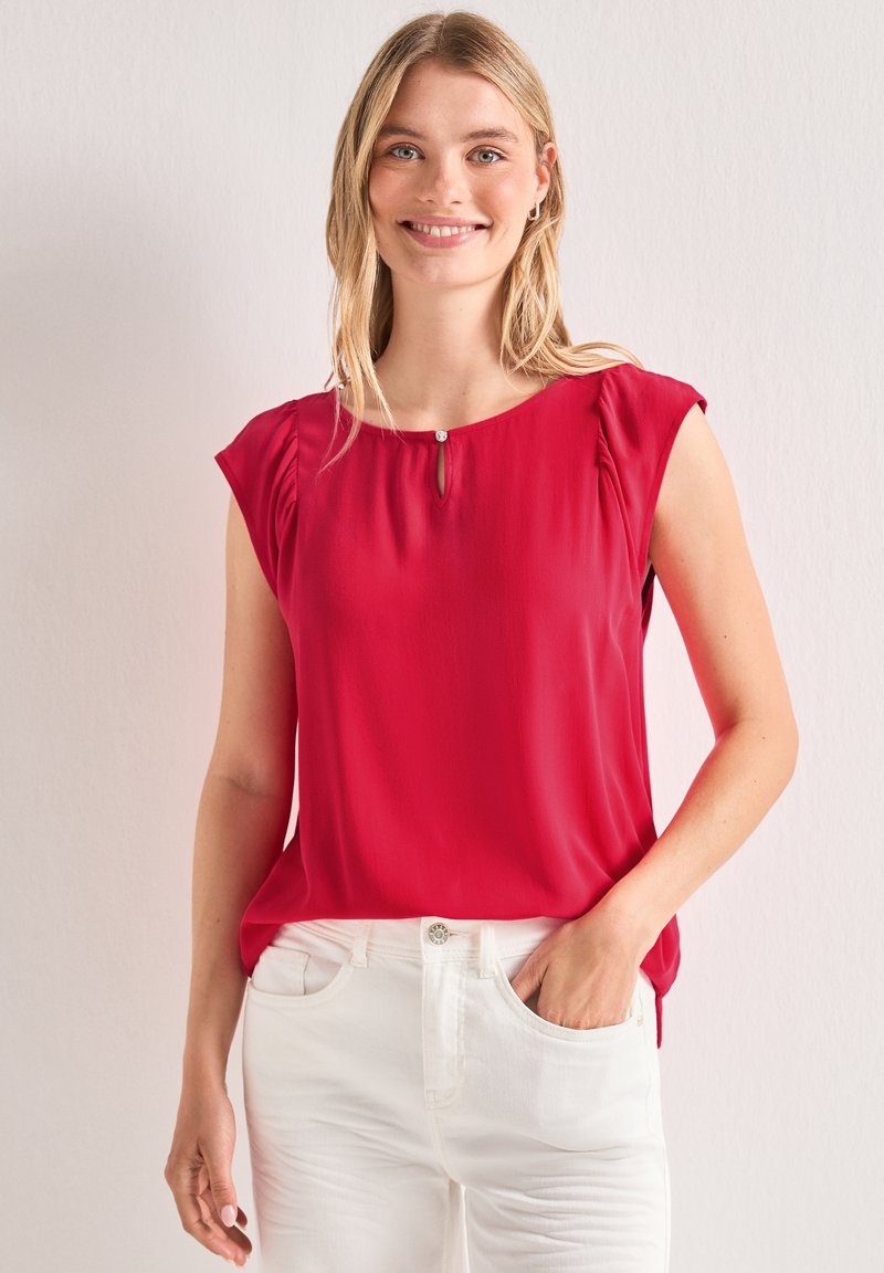 Femme blonde souriante portant un chemisier rouge sans manches et un pantalon blanc, une main dans la poche, debout devant un fond uni.