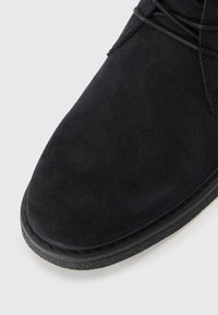 Botín de ante negro con punta redonda, cordones mínimos y suela de goma negra texturizada. Diseño limpio con un acabado de material suave.