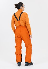 Salopettes imperméables orange avec bretelles noires ajustables et fermetures éclair aux chevilles. Dotées d'une taille élastique pour le confort et la liberté de mouvement.