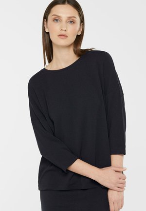 Woman with straight brown hair wearing a loose-fitting black long-sleeve top, standing with arms crossed gently in front.