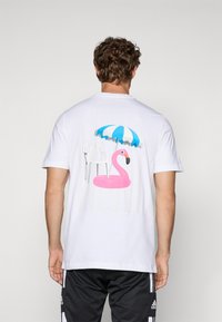 Hvid T-shirt med et farverigt grafik: en pink flamingo, en hvid stol og en blå-hvid stribet parasol på ryggen.