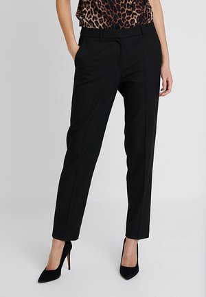 Pantalones negros de corte ajustado, con una textura suave, frente plano y dos bolsillos laterales. Llevados con zapatos de tacón alto negros.