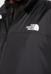 Gros plan d'une personne portant une veste noire North Face avec un logo de marque visible sur la poitrine et une couche intérieure noire partiellement zippée.