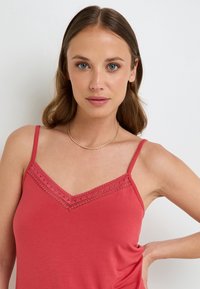 Camisole rouge avec des fines bretelles réglables, ornée d'une dentelle le long du décolleté en V. Le tissu semble doux et lisse.