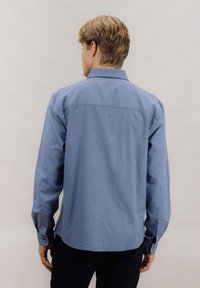 Lys blå, langermet skjorte laget av stoff med glatt tekstur. Har standard krage og knappede mansjetter. Rett kant nederst.