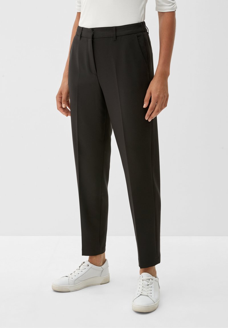 Personne portant un pantalon noir sur mesure longueur cheville avec pince devant, associé à des baskets blanches et un haut blanc, debout sur un fond uni.
