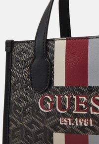 Mustat ja kuviolliset kangaskassit, joissa on nahkareunat, punaisia, valkoisia ja harmaita vaakaraitoja sekä "GUESS"-brändäys paksuilla kirjaimilla.