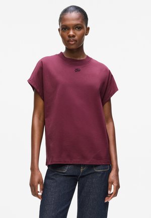 Maroon cotton t-shirt with short sleeves, crew neck, and small black logo on the chest. Smooth texture, relaxed fit, paired with dark jeans.