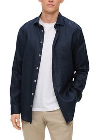 Camicia a maniche lunghe in lino blu navy con colletto, chiusura a bottoni e bottoni bianchi, indossata sopra una t-shirt bianca, abbinata a pantaloni beige chiaro.